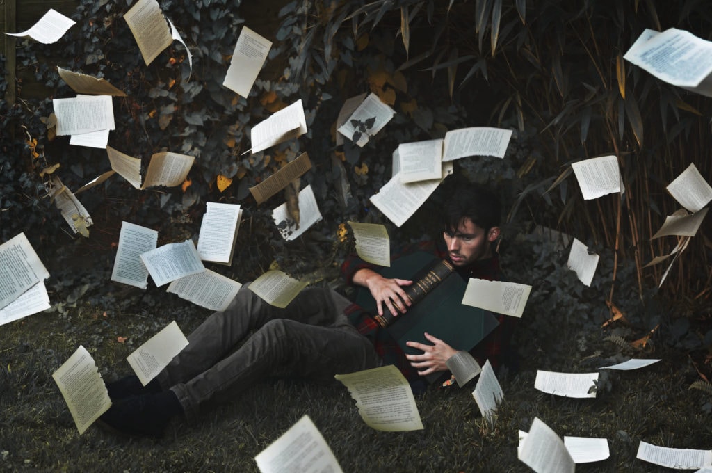 Chico sentado en la hierba con libro y muchas hojas del libro volando