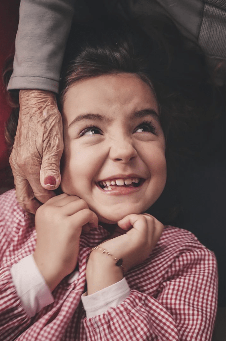 Niña sonriendo mientras la mano de la yaya le hace cosquillas en el cuello, ganadora del #fotoreto87 con el tema ternura