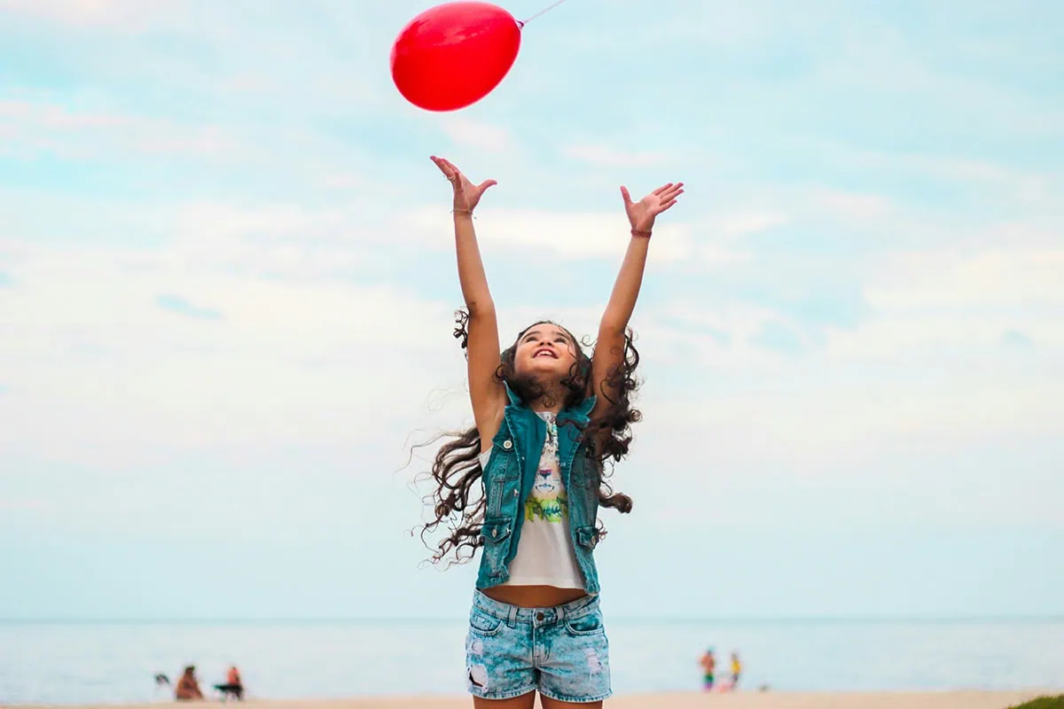 Niña saltando y lanzando un globo rojo al cielo en la playa, uso back button focus